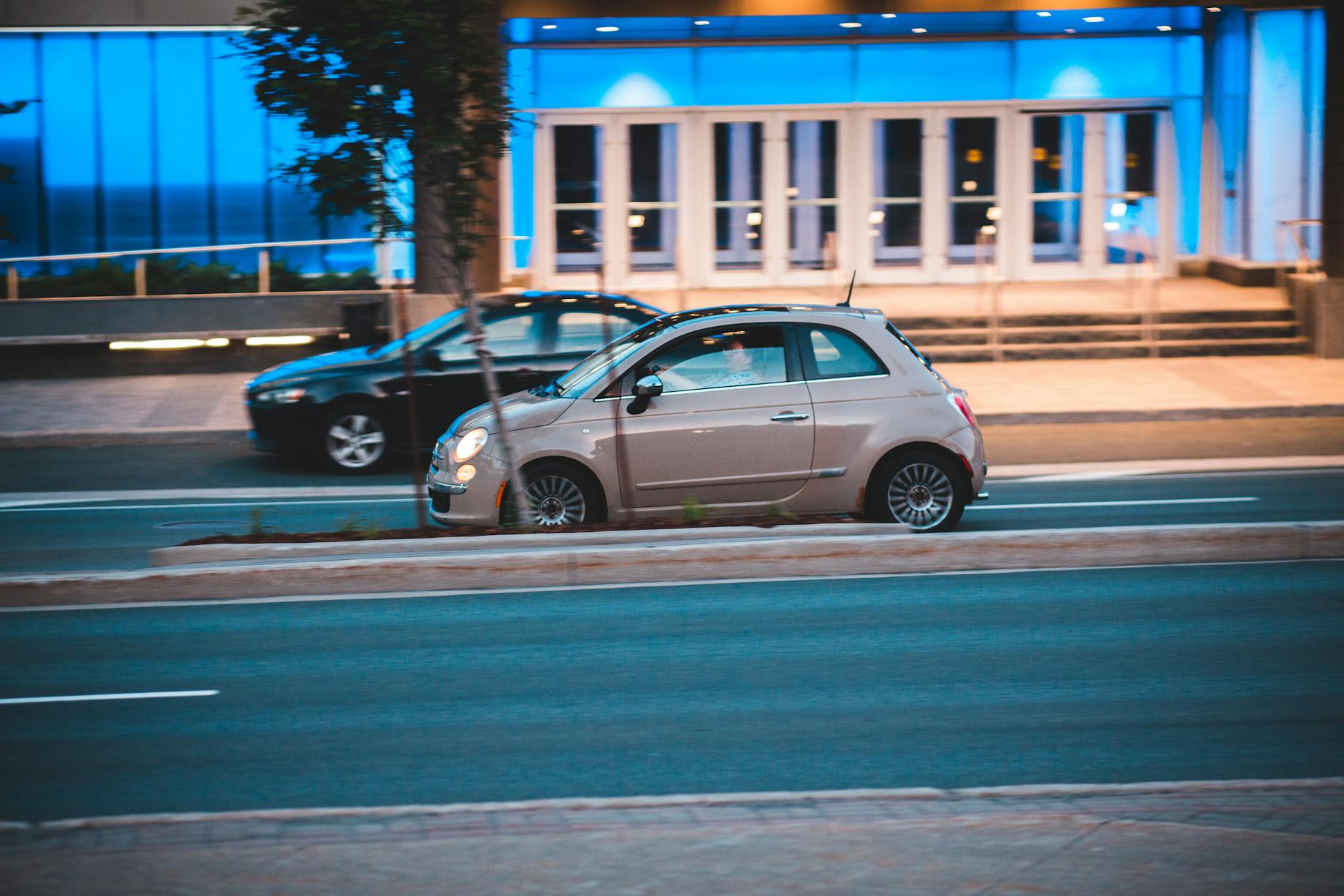 Fiat 500 jadący nocą ulicą obok nowoczesnego, podświetlonego budynku