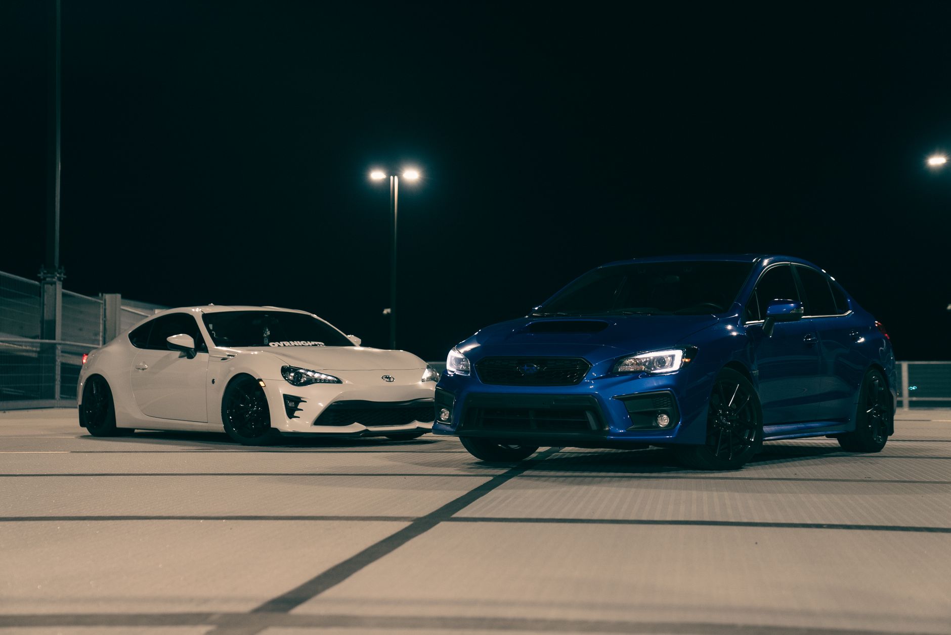 Dwie sportowe Hondy Civic, biała i niebieska, na dachu parkingu nocą Dwie sportowe Hondy Civic, biała i niebieska, na dachu parkingu nocą